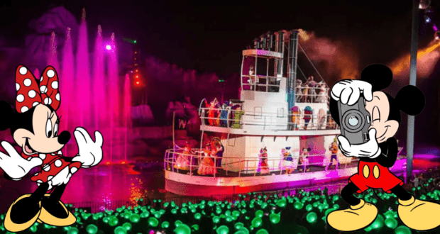 Mickey Mouse snaps a photo of Minnie Mouse as the vibrant Fantasmic! nighttime boat show illuminates the water, with cheering guests glowing in the audience behind them at Disney World.