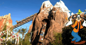 Goofy stands looking puzzled in front of Expedition Everest at Disney’s Animal Kingdom, as guests speed by on the snowy mountain roller coaster.
