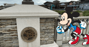 Mickey Mouse dashes past a stone post adorned with a Disney Springs plaque, embodying the excitement of runDisney festivities as lively shops and happy guests create a bustling backdrop.