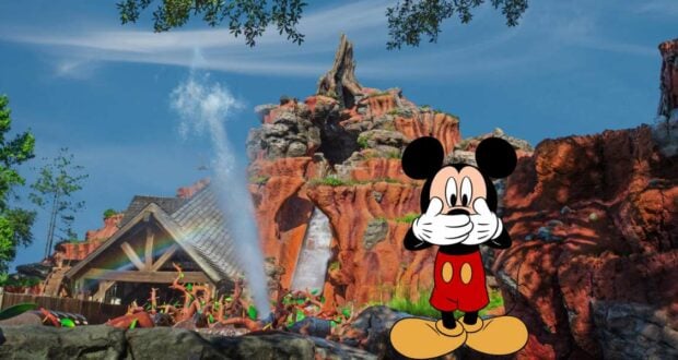 Mickey Mouse playfully covers his mouth while posing in front of Splash Mountain at Disney World, where a thrilling splash sends water soaring beneath the sunny blue sky.