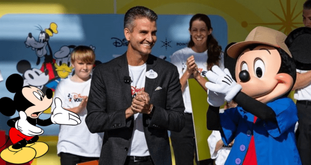 A Disney executive in a suit enthusiastically claps alongside Mickey Mouse, surrounded by happy fans in Disney shirts. The scene brims with the signature excitement and magic found at Disney World, echoing the inspiring energy of a Josh D'Amaro interview.