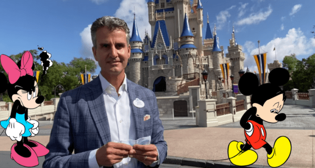 A guest poses in front of Cinderella Castle at Disney World, flanked by sad-looking cartoon Minnie and Mickey Mouse, both holding a sign referencing Josh D’Amaro and Disney’s annual pass program.