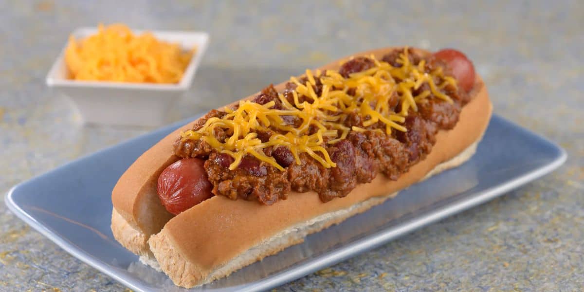 A chili cheese hot dog on a plate, smothered with ground beef chili and topped with shredded cheddar. In the background, a small bowl of grated cheese adds to this hearty meal's allure, making it one of Disney Parks' ten satisfying on-the-go delights.