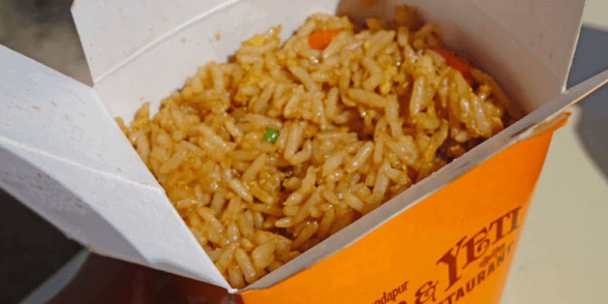 A close-up of a white takeout box reveals fried rice mixed with small veggies, an ideal choice from the Ten Hearty On the Go Meals at Disney Parks. The box's orange exterior features partial text, hinting at its delightful contents.