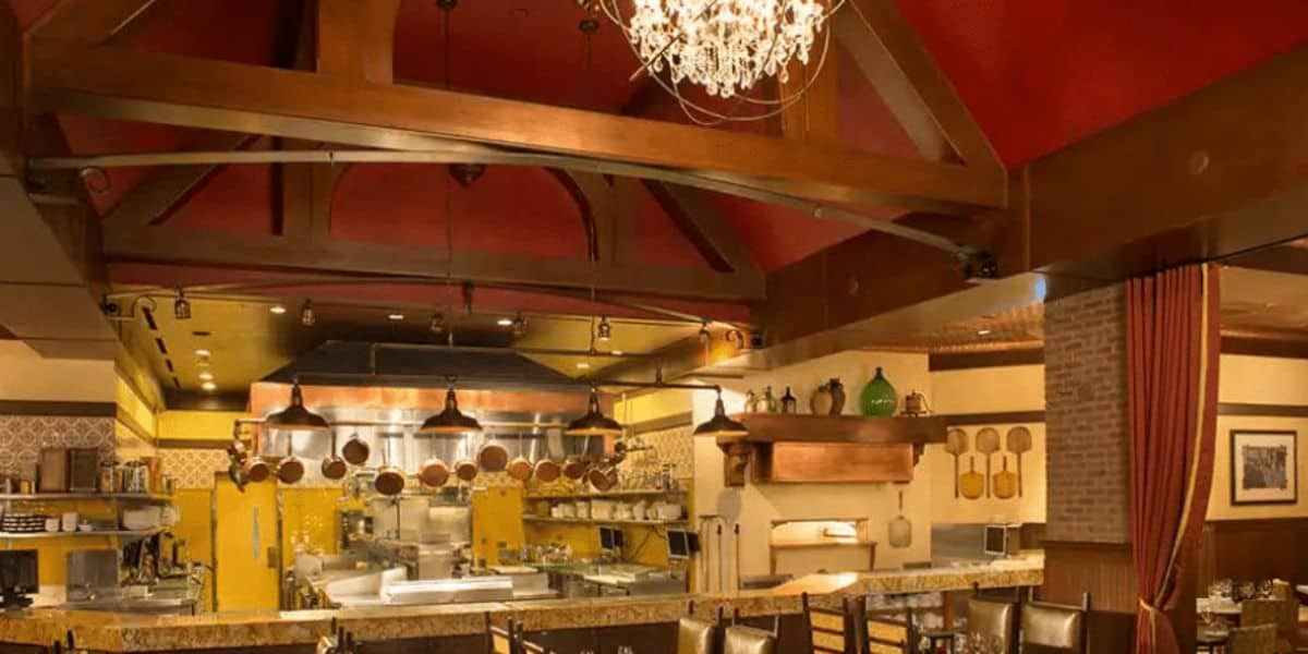 The interior of this cozy restaurant near Walt Disney World boasts a warm ambiance, highlighted by a high, wooden beamed ceiling and a chandelier. An open kitchen with visible cooking areas invites guests to enjoy the aroma of garlic bread, while rustic decor and earth-toned walls enhance the charm.