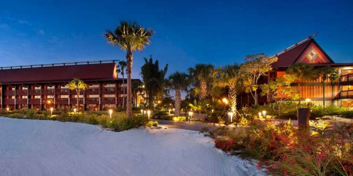 Nighttime view of a tropical resort reminiscent of the Disney Vacation Club Polynesian, with red-roofed buildings and warm lighting. Surrounded by palm trees and lush greenery, a white sandy area graces the foreground. The deep blue sky suggests dusk or early evening.