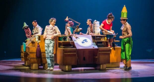 Cirque du Soleil Drawn to Life performers dazzle the stage in vibrant costumes, energizing the audience as they bring Disney animation to life around a grand wooden desk beneath theatrical spotlights.