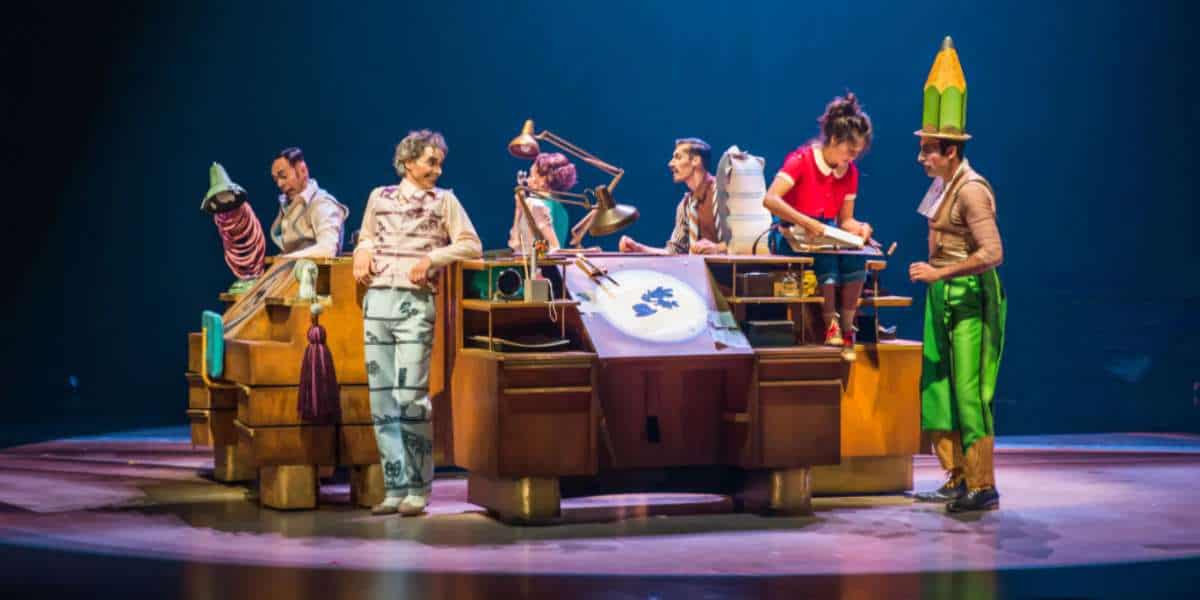 Cirque du Soleil Drawn to Life performers dazzle the stage in vibrant costumes, energizing the audience as they bring Disney animation to life around a grand wooden desk beneath theatrical spotlights.