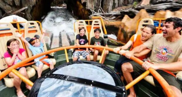 A group of people, including children and adults, are on the Kali River Rapids ride, holding onto the orange safety bars. They are smiling and laughing as they face an upcoming splash of water, set against a rocky backdrop.