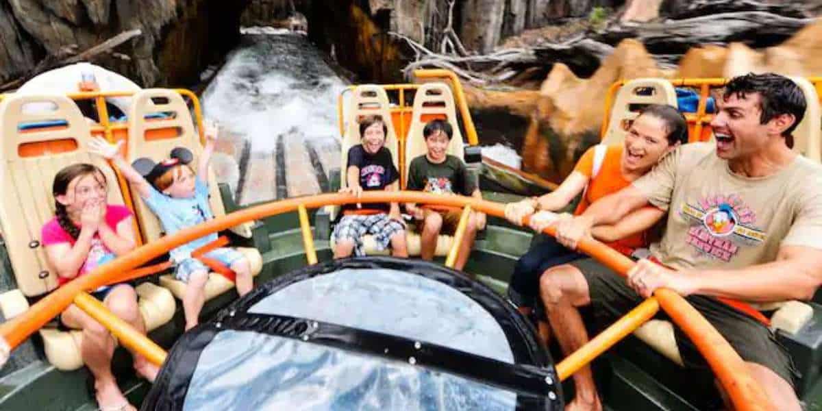 A group of people, including children and adults, are on the Kali River Rapids ride, holding onto the orange safety bars. They are smiling and laughing as they face an upcoming splash of water, set against a rocky backdrop.