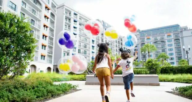 Two kids dash along a path outside a sleek apartment-style Disney Resort, each clutching a Mickey Mouse balloon, their joy mirroring the thrill of exploring all the family-friendly options at Walt Disney World.