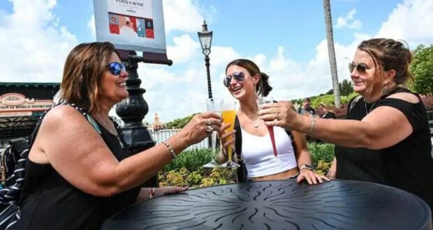 Three friends clink wine glasses and share smiles at an outdoor table in Walt Disney World, enjoying a sunny day and the magic of relaxation and camaraderie at one of the resort’s best spots for wine lovers.