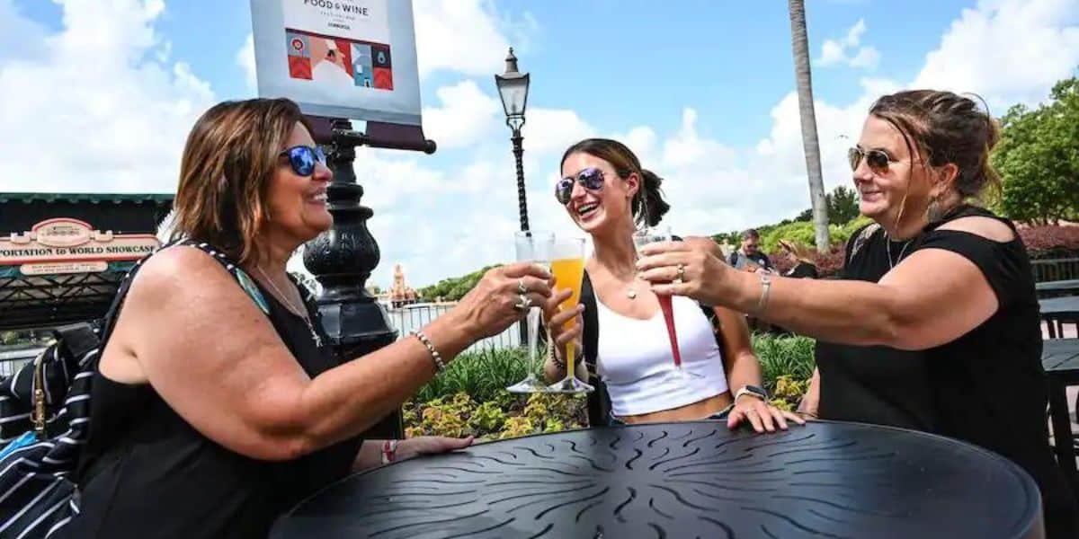 Three friends clink wine glasses and share smiles at an outdoor table in Walt Disney World, enjoying a sunny day and the magic of relaxation and camaraderie at one of the resort’s best spots for wine lovers.