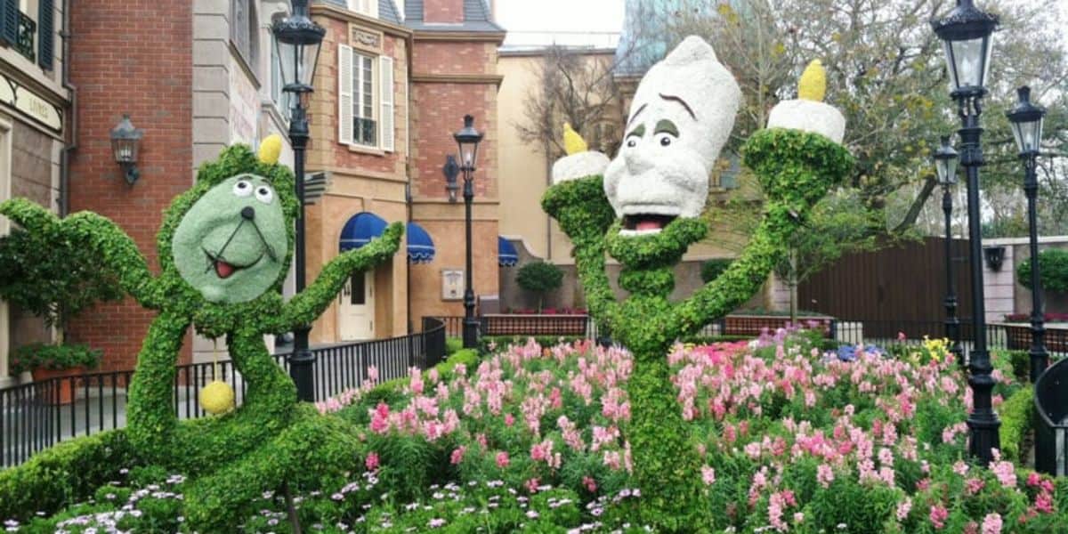 Two whimsical topiary figures from Disney's "Beauty and the Beast"—Lumière and Cogsworth—stand decorated with green foliage and flowers. Surrounded by a colorful garden filled with pink and white blooms, these charming displays are one of the must-dos at Flower & Garden Festival, complemented by quaint buildings and lampposts.