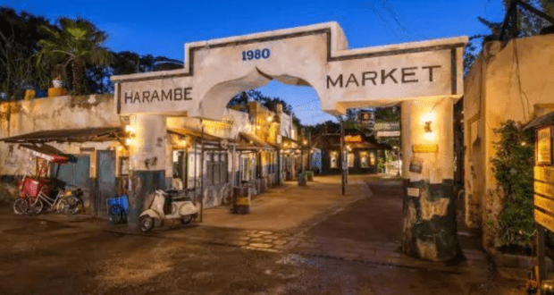 Entrance to Harambe Market, a rustic marketplace with an arch displaying "1980" and "Harambe Market." The scene reveals a cobblestone path, streetlights, scooters, and food carts under a twilight sky. Whispers of secret passages at Disney World add an air of mystery to this enchanting setting.