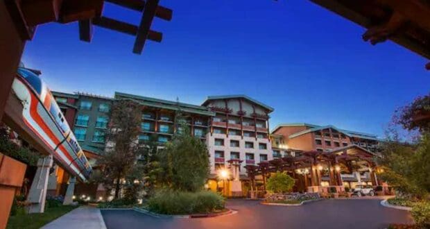 A modern hotel with warm lighting is seen at dusk, surrounded by greenery. A monorail glides by on an elevated track beside the building, its windows reflecting the deep blue sky—inside, Disney Resort Soap Dispensers add a whimsical touch.