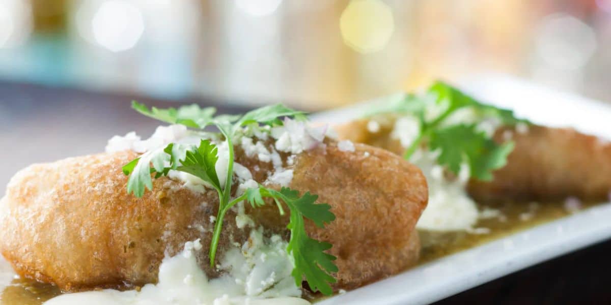 Close-up of a plated dish featuring two stuffed peppers topped with creamy sauce, crumbled cheese, and fresh cilantro. Dine inside this popular Disney ride as the vibrant dish is presented on a rectangular white plate with a blurred background, adding to the enchanting experience.