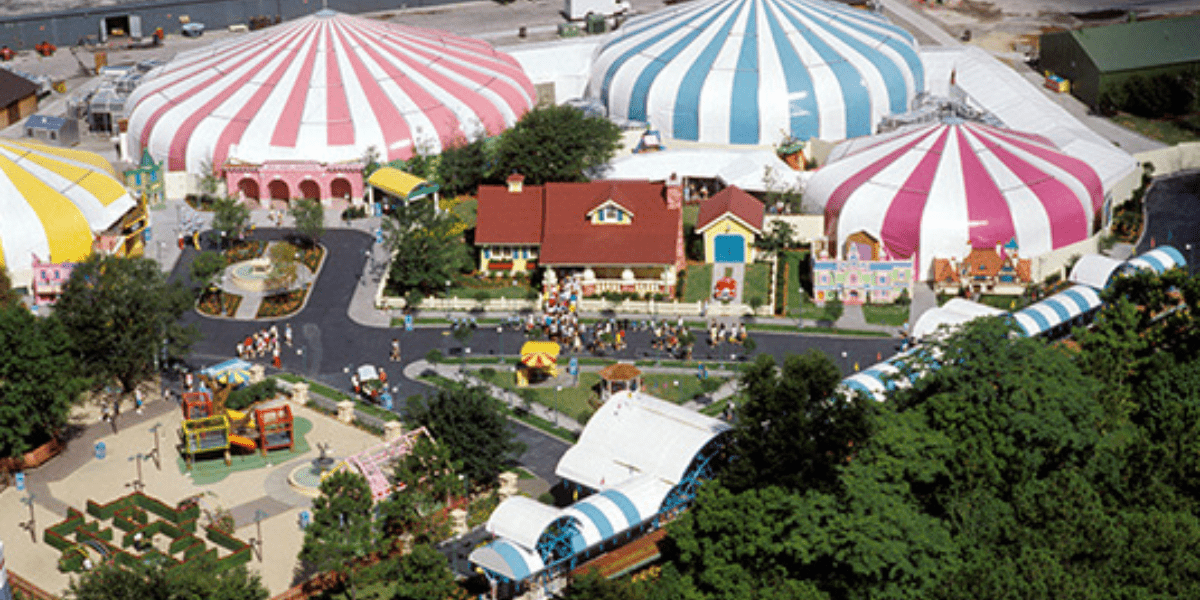 Aerial view of a colorful amusement park reminiscent of Mickey's Toontown at Disney World, with pink, blue, and yellow striped circus tents. The park features winding pathways, charming small buildings, and green trees scattered throughout as visitors explore the lively attractions.