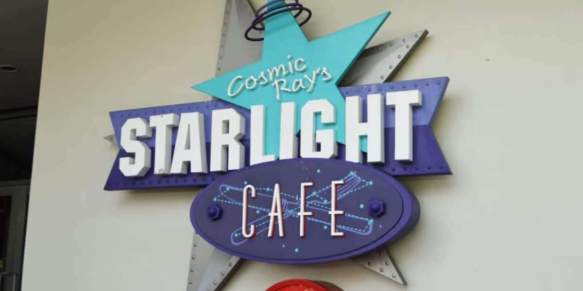 A colorful sign for "Cosmic Ray's Starlight Cafe" at Disney World. The sign features a large star with teal and purple accents and illuminated white text. A smaller oval below has "Cafe" in neon-style letters, with a rocket graphic, against a white wall background—a true gem for those who know the Disney World Secret Menu.