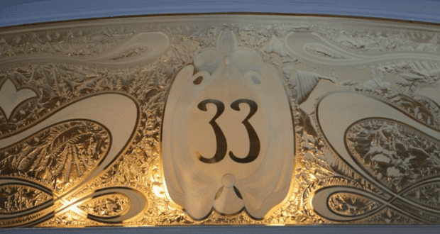 An ornate plaque showcasing the number "33" is embellished with intricate gold and cream swirling designs. The background features detailed leaf-like motifs softly lit, reminiscent of the exclusive allure associated with off-limits locations at Disney Parks.
