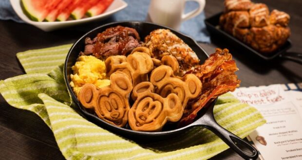 A skillet filled with Mickey Mouse-shaped waffles, scrambled eggs, bacon, and sausage sits on a green striped cloth at Garden Grill Restaurant. In the background, there's watermelon, a pastry, and a small pitcher of milk, with a Disney menu partially visible on the side.