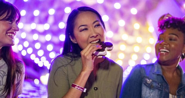 Three women smile and laugh under string lights, with one woman in the center holding a large s’more—sharing tips from their Disney vacation packing list in a warmly lit, festive atmosphere.