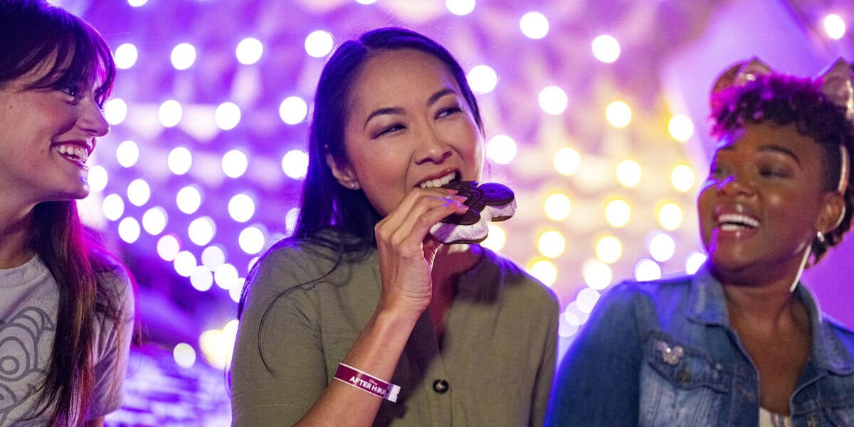 Three women smile and laugh under string lights, with one woman in the center holding a large s’more—sharing tips from their Disney vacation packing list in a warmly lit, festive atmosphere.