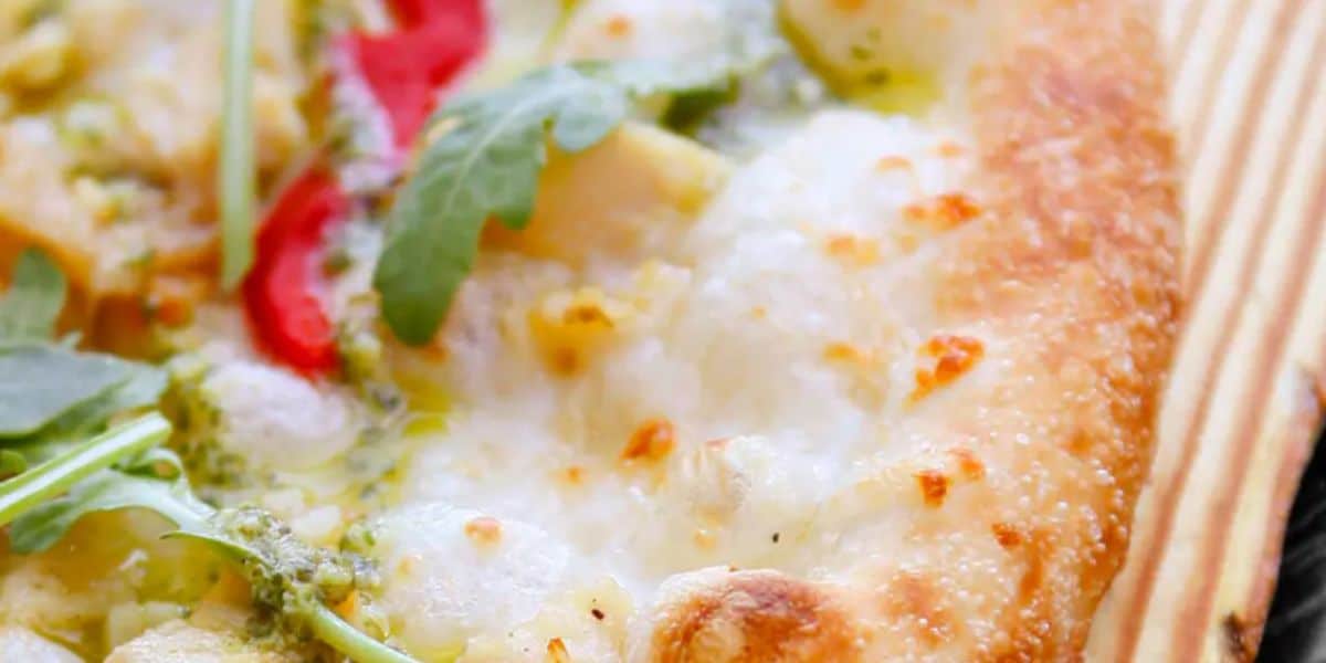 Close-up of a pizza in one of the ten dining recommendations for your Disney vacay, featuring a golden-brown crust, melted cheese, sliced red peppers, vibrant green arugula leaves, and a drizzle of green pesto sauce on top.