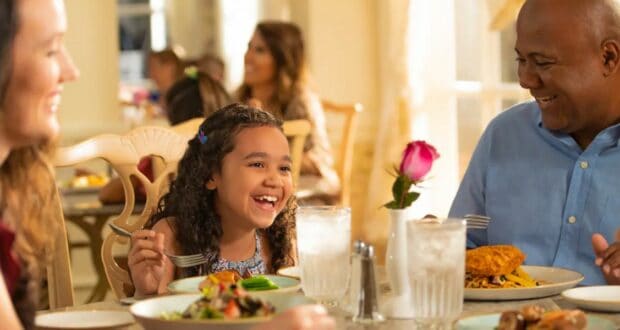 A smiling family enjoys a meal reminiscent of our favorite moderately priced restaurants at Walt Disney World. A young girl laughs while holding a fork, surrounded by plates of food and glasses of water. A man in a blue shirt smiles at her, as a pink rose in a vase graces the table.
