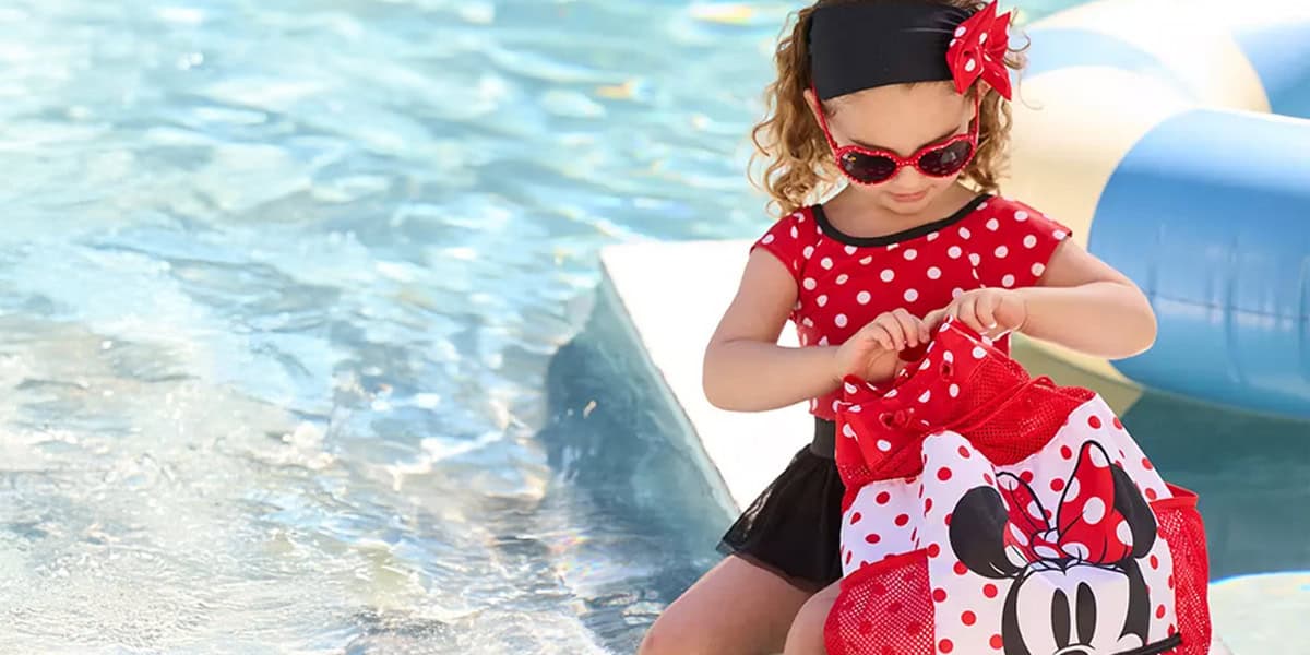 A young girl in a red polka-dot dress, headband, and sunglasses sits by a pool, opening a Minnie Mouse bag—perfect for any Disney vacation packing list. An inflatable pool float is visible in the background.