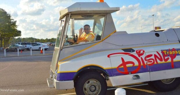 A cheerful Disney Cast Member in a safety vest waves from a classic Disney Transport tram, just like the ones that shuttle guests to the gates of EPCOT and Hollywood Studios, basking in the sunshine of a bustling theme park parking lot.