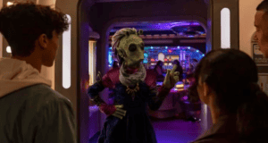 A group of people stand at the entrance of a futuristic room reminiscent of Star Wars: Galactic Starcruiser. An extraterrestrial figure dressed in colorful attire gesticulates while facing them, set against a backdrop of vibrant lights and sleek, sci-fi interiors.