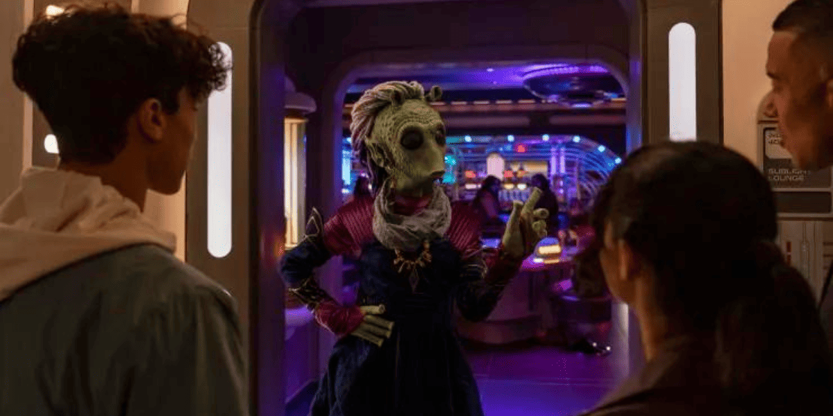 A group of people stand at the entrance of a futuristic room reminiscent of Star Wars: Galactic Starcruiser. An extraterrestrial figure dressed in colorful attire gesticulates while facing them, set against a backdrop of vibrant lights and sleek, sci-fi interiors.