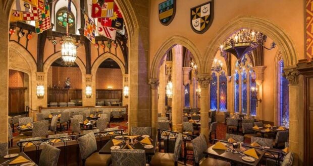 An elegant, medieval-themed dining hall in the heart of Magic Kingdom park features arched stone doorways, numerous chandeliers, and colorful flags hanging from the ceiling. Tables are set with yellow napkins and cushioned seating, creating a warm, inviting atmosphere for the best dining experience.