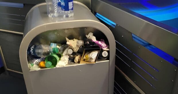 An overflowing trash bin with assorted waste, including plastic bottles, paper, and food packaging. A plastic water bottle rests on top. Situated in a modern, metallic setting with blue lighting, it highlights the potential for Disneyland recycling initiatives to make a difference.