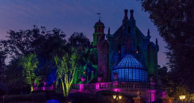 A spooky, dark mansion is lit with eerie green, purple, and blue lights against a twilight sky. Surrounded by trees, the building evokes Disney's Haunted Mansion ride with its distinct glass conservatory, towers, and chimney stacks—perfect for a haunted mansion trivia night.