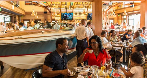 A lively restaurant scene with a boat displayed indoors sets the stage for romance at Disney Springs. Diners savor meals, engaging in conversation amidst a bustling atmosphere. Servers in crisp white shirts attend to guests, while hanging lights and TVs enhance the charming ambiance.