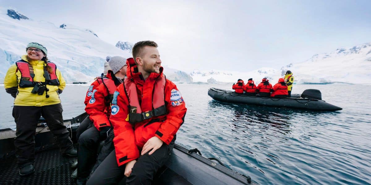 A group wearing red jackets and life vests sits on a boat in icy waters with snowy mountains looming in the background. Nearby, another boat mirrors this adventure. Under an overcast sky, this remote setting invites you to experience new vacations by Disney in a breathtaking winter wonderland.