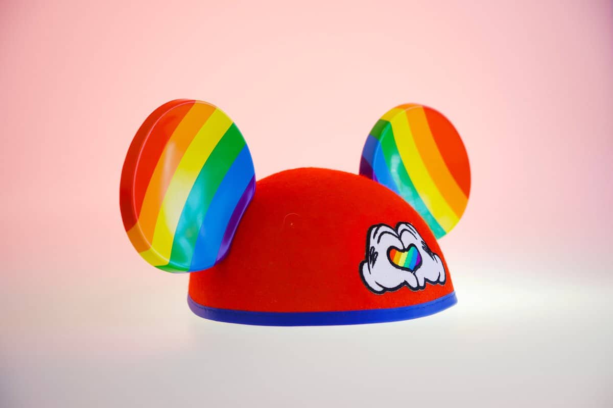 Colorful Mickey Mouse ears featuring vibrant rainbow stripes and a heart-shaped hand patch, set against a playful pink gradient—an iconic accessory beloved by Disney Parks guests and perfect for showing your pride on your next visit to Disney World or Disneyland.