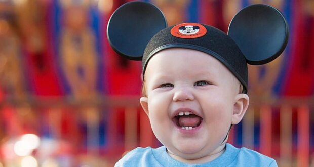 A baby wearing Mickey Mouse ears is smiling joyfully, capturing the magic of visiting Disney's EPCOT with a toddler. The colorful and blurred background evokes an amusement park setting, perfect for exploring Disney tips for moms on making memories with their little ones.