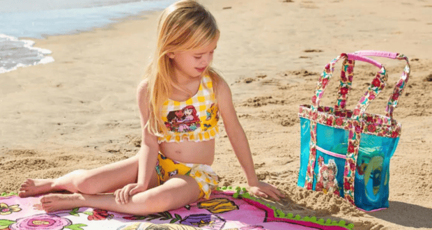 A young guest in a bright yellow swimsuit relaxes on a beach towel by the seaside, with a vibrant Disney merchandise bag set on the sand close by.