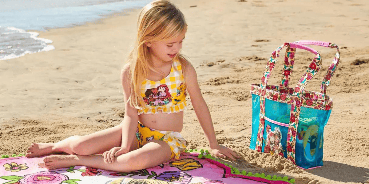 A young guest in a bright yellow swimsuit relaxes on a beach towel by the seaside, with a vibrant Disney merchandise bag set on the sand close by.