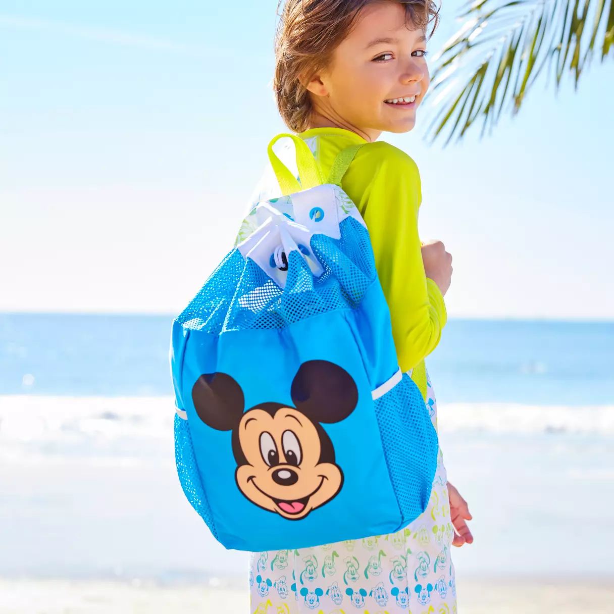 Young Disney fan sporting a blue Mickey Mouse backpack enjoys a sunny day by the ocean, capturing the magic and playful spirit of Disney merch outside the parks.