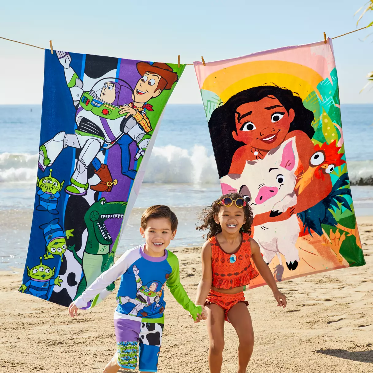 Two excited kids in swimwear hold hands on the beach, with vibrant Toy Story and Moana Disney towels hanging behind them—showcasing beloved Disney characters and magical merchandise for young fans.