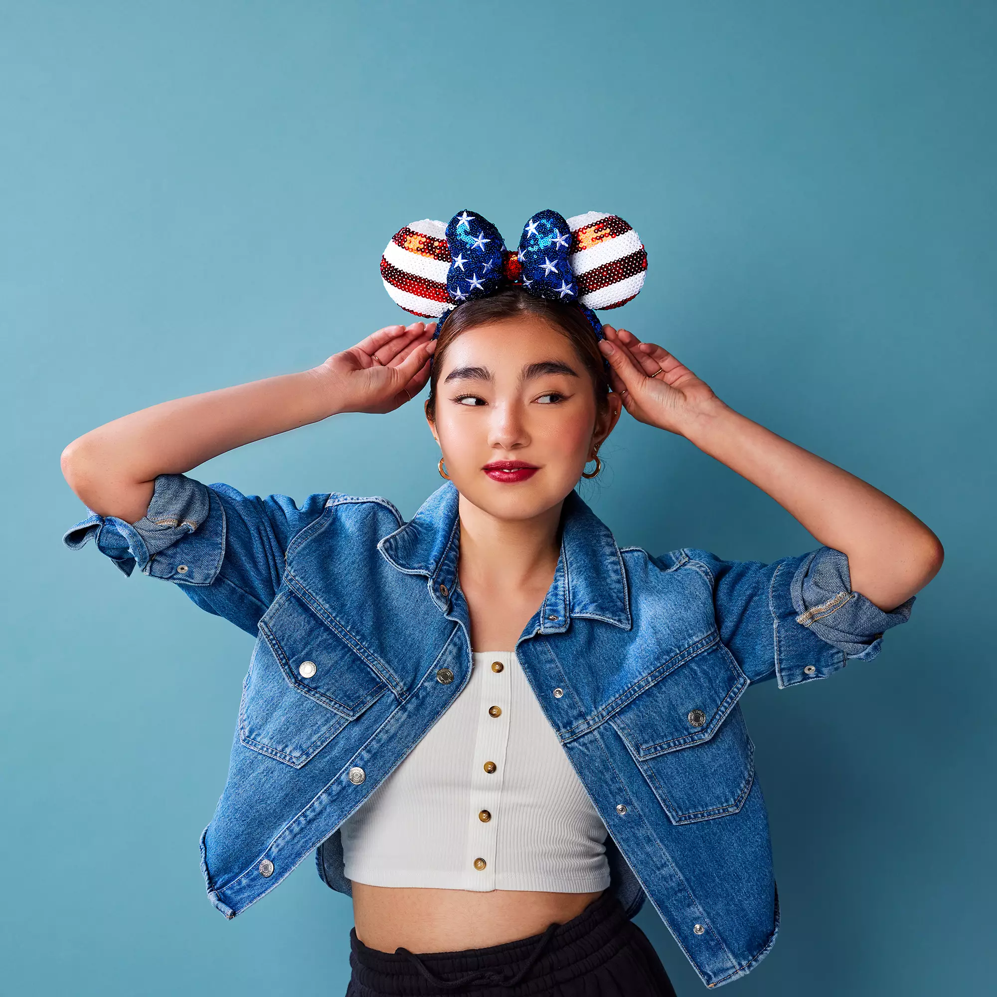 Disney fan sports a denim jacket and star-spangled Minnie Mouse ears in front of a vibrant blue backdrop, proudly showing off her park-ready style.