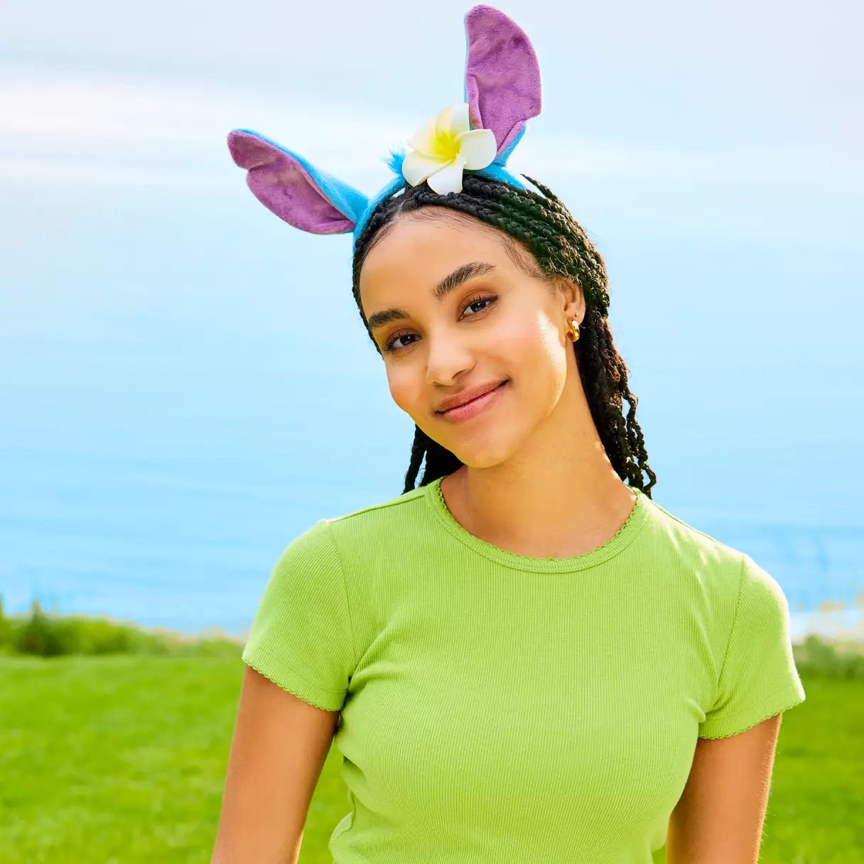 A Disney fan beams in a vibrant green shirt outside, sporting a playful headband with purple ears and a white flower—an ideal accessory for showing off her passion for Disney parks merchandise.