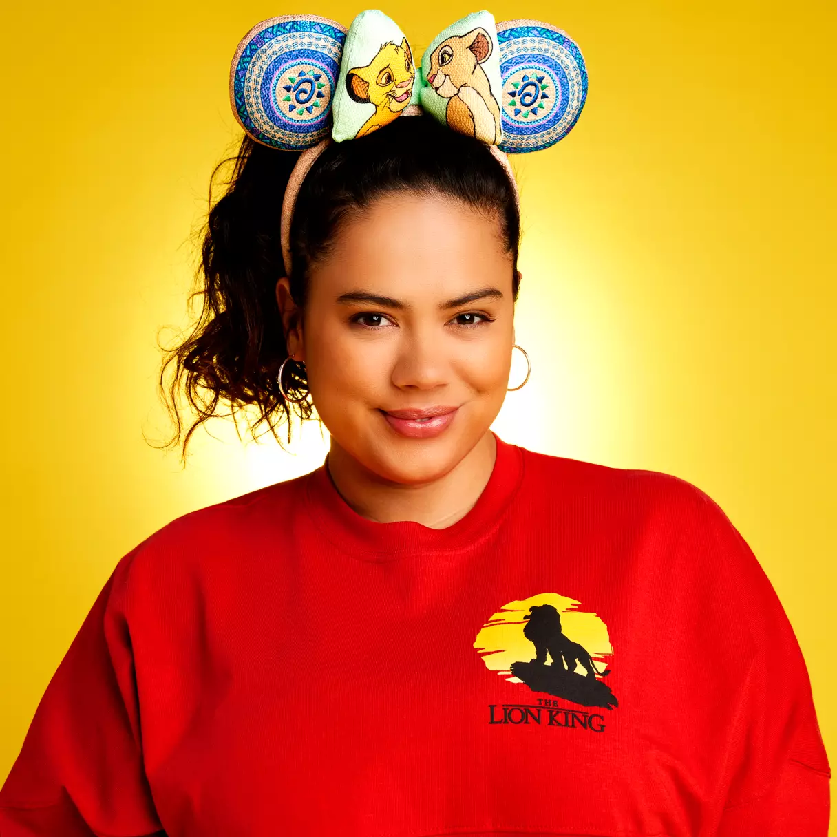 Disney fan sporting a red Lion King shirt and Simba ear headband, beaming with her favorite merch against a bold yellow Disney-inspired backdrop.