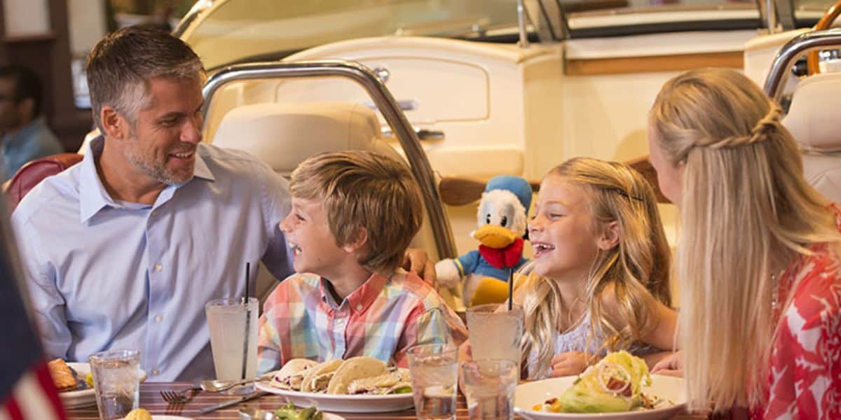 A family of four, including two children, enjoys a meal at a restaurant. They are smiling and sitting at a table with food and drinks. A plush toy resembling a cartoon duck is visible in the background, hinting at the delightful dining options at Disney World.