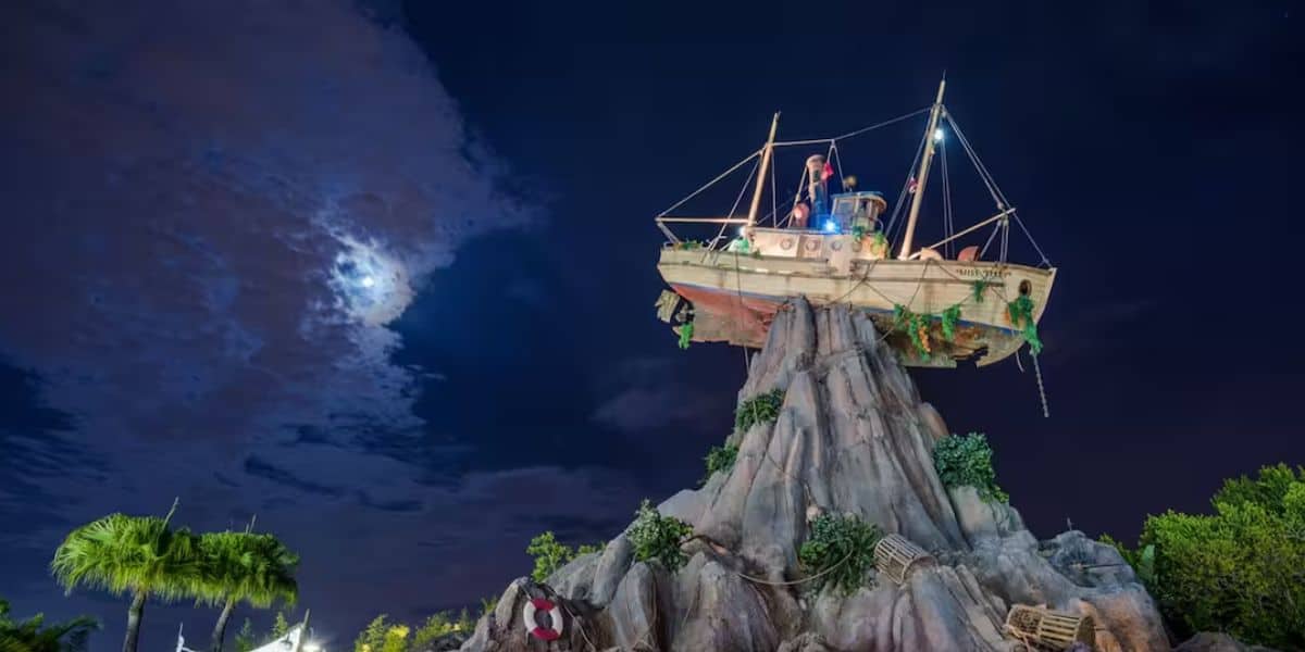 A dramatic night sky with a partially cloud-covered moon illuminates a ship structure perched atop a rocky formation, reminiscent of Disney Typhoon Lagoon. Palm trees and scattered wooden crates decorate the scene below, evoking the adventure of a volcanic island.