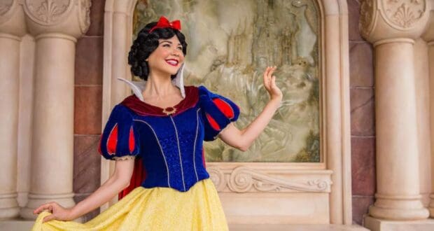A person dressed as a fairytale character stands in front of a decorative backdrop featuring a castle at Disney World Princess Week. They wear a red bow, blue and red top, and yellow skirt, posing with one hand raised. The setting is ornate with stonework.
